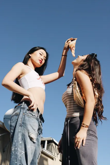 Two women against a blue sky, one feeding the other a slice of pizza