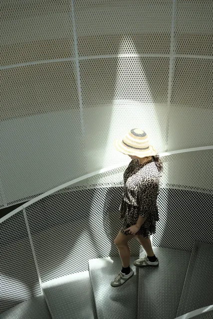 A woman in a hat on a perforated metal spiral staircase under a skylight